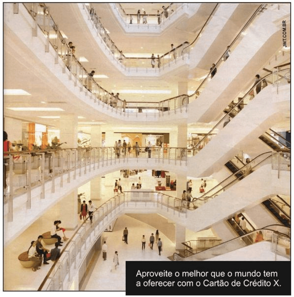 Anúncio com interior de shopping, escadas rolantes e pessoas. Texto: Aproveite o melhor que o mundo tem a oferecer com o Cartão de Crédito X. A identificação vertical do crédito fotográfico está preservada na imagem.